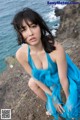 A woman in a blue dress sitting on a rock by the ocean.